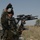A U.S. Army soldier in ACU (Army Combat Uniform) pattern camouflage is giving a thumbs-up hand signal while holding an M4 carbine with an M203 grenade launcher attachment. The soldier is equipped with full combat gear including a helmet, tactical vest, and gloves. The background suggests an arid, Middle Eastern environment with sparse vegetation, palm trees, and a dilapidated structure.