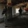Two armed soldiers in U.S. Army digital camouflage (ACU pattern) are conducting a clearing operation inside a dilapidated, abandoned building. The interior shows significant deterioration with damaged walls, dirt floors, and barred windows. Hanging from the ceiling are what appear to be inverted plastic poultry feeders, suggesting the structure was previously used as an agricultural or livestock facility.