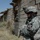 A U.S. Army soldier in full combat gear is positioned against the exterior wall of a damaged concrete block building in an arid environment. The soldier is wearing Army Combat Uniform (ACU) digital camouflage and bears a 2nd Infantry Division shoulder patch. The setting appears to be a rural or semi-urban area consistent with Middle Eastern terrain.