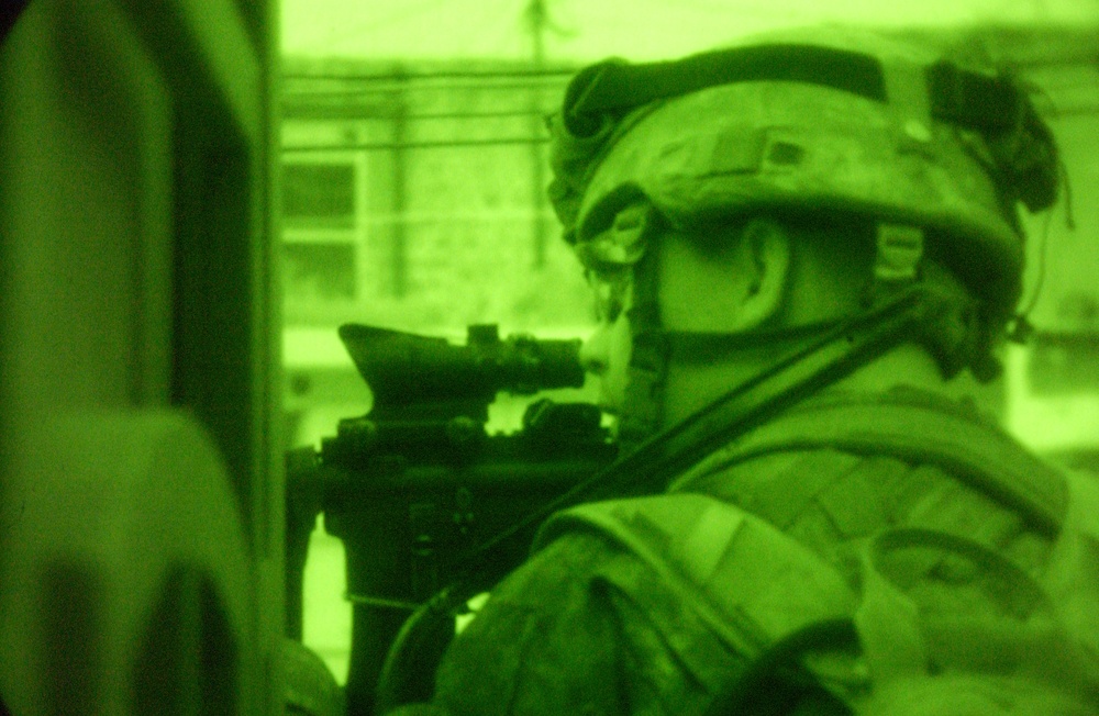 A night-vision captured image showing a soldier in full combat gear aiming a rifle with an optical scope. The image is rendered entirely in green monochrome, characteristic of night-vision device (NVD) photography. The soldier appears to be in an urban environment, positioned near a doorway or wall.