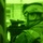 A night-vision captured image showing a soldier in full combat gear aiming a rifle with an optical scope. The image is rendered entirely in green monochrome, characteristic of night-vision device (NVD) photography. The soldier appears to be in an urban environment, positioned near a doorway or wall.