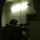A heavily equipped soldier in full combat gear is photographed from behind in a low-light interior corridor, appearing to be conducting a room-clearing or building-entry operation. The soldier is raising a leg in a kicking motion toward a closed door while aiming a rifle with an optical scope. The image is taken through an arched doorway, suggesting the photographer is positioned in a trailing or overwatch position.