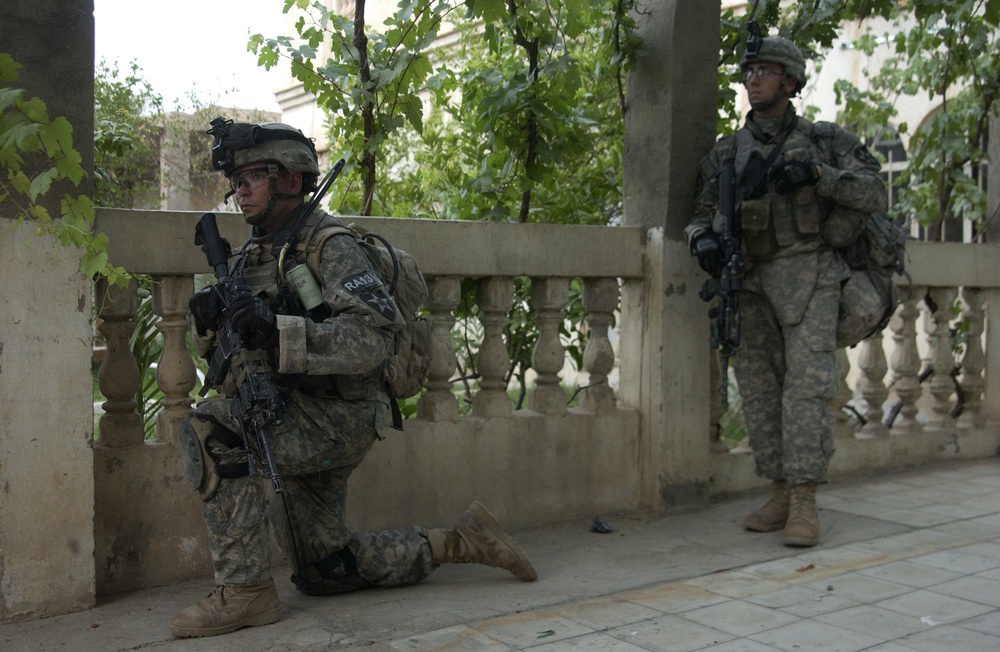Two U.S. Army soldiers in full combat gear are positioned along a decorative stone balustrade in what appears to be an urban environment with trees and buildings visible in the background. The soldier in the foreground is kneeling in a tactical position while holding an assault rifle, while the second soldier stands alert nearby. The setting and equipment suggest a combat patrol or security operation in a Middle Eastern urban area.