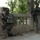 Two U.S. Army soldiers in full combat gear are positioned along a decorative stone balustrade in what appears to be an urban environment with trees and buildings visible in the background. The soldier in the foreground is kneeling in a tactical position while holding an assault rifle, while the second soldier stands alert nearby. The setting and equipment suggest a combat patrol or security operation in a Middle Eastern urban area.