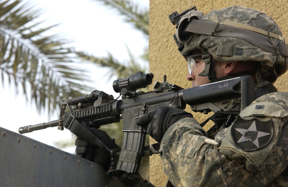 A U.S. Army soldier in full combat gear is shown in a tactical aiming position with an M4 carbine rifle. The soldier wears Army Combat Uniform (ACU) digital camouflage and displays a 2nd Infantry Division shoulder patch. The setting appears to be an urban or semi-arid environment, consistent with Middle Eastern deployment.