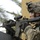 A U.S. Army soldier in full combat gear is shown in a tactical aiming position with an M4 carbine rifle. The soldier wears Army Combat Uniform (ACU) digital camouflage and displays a 2nd Infantry Division shoulder patch. The setting appears to be an urban or semi-arid environment, consistent with Middle Eastern deployment.