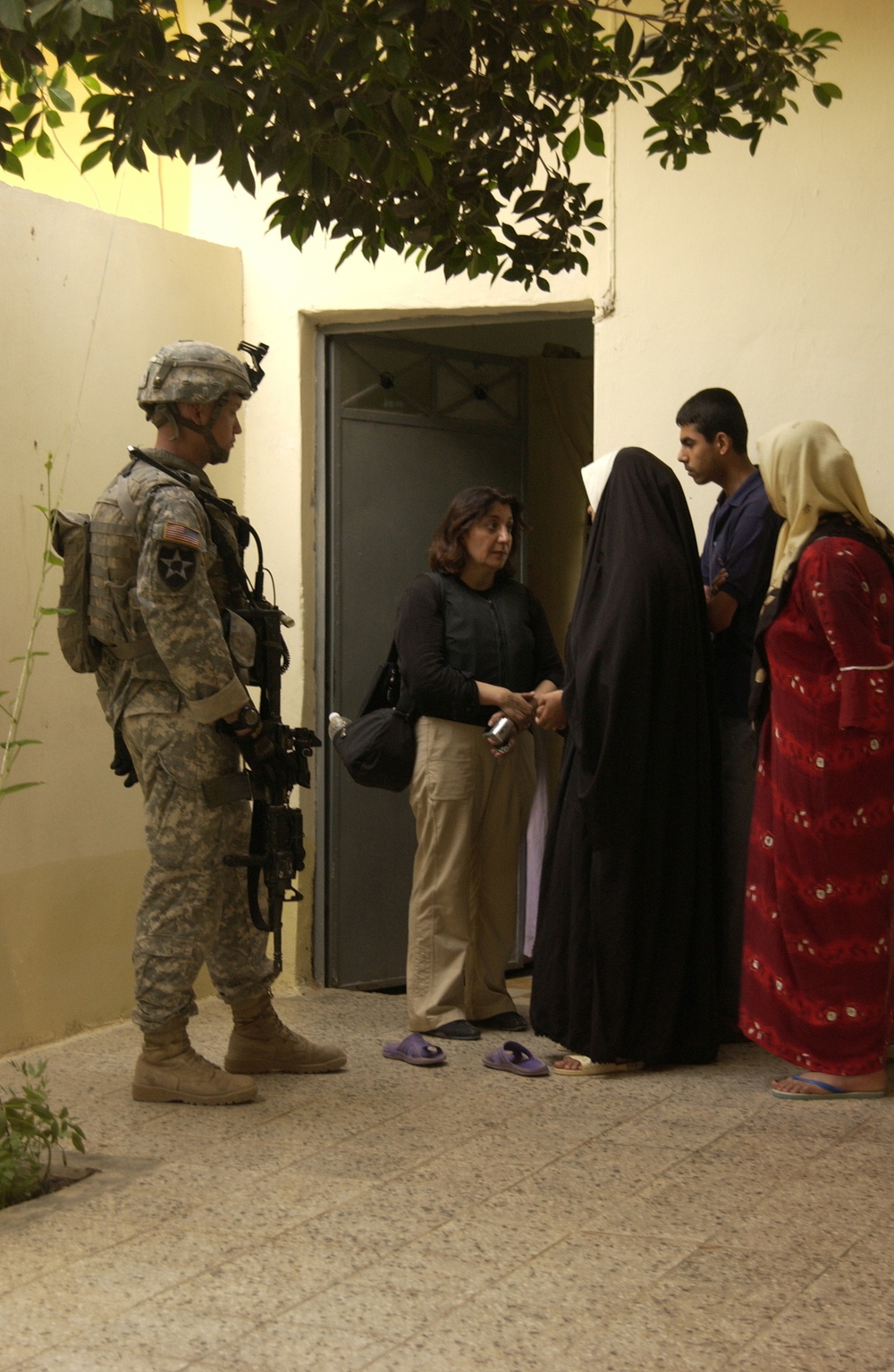 A U.S. Army soldier in full combat gear stands near a doorway while a civilian woman, likely an interpreter or civil affairs personnel, engages in conversation with a group of local residents including a woman in a black abaya, a young man, and a woman in a red dress with a head covering. The setting appears to be a residential courtyard or building entrance, suggesting a community engagement or house visit operation. The scene is lit by ambient artificial light, indicating an evening or nighttime interaction.
