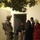 A U.S. Army soldier in full combat gear stands near a doorway while a civilian woman, likely an interpreter or civil affairs personnel, engages in conversation with a group of local residents including a woman in a black abaya, a young man, and a woman in a red dress with a head covering. The setting appears to be a residential courtyard or building entrance, suggesting a community engagement or house visit operation. The scene is lit by ambient artificial light, indicating an evening or nighttime interaction.
