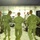 Military personnel in camouflage uniforms are dancing or moving energetically in front of a live band performing on a small stage inside what appears to be a military facility or tent structure. The scene depicts an informal morale event or entertainment gathering for troops. A live band with guitars and microphones performs while soldiers enjoy the music in the foreground.