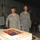 Two U.S. Army soldiers are cutting a large sheet cake decorated with an American flag design, likely at a military celebration or ceremony. The soldier on the left, identified by his name tape as 'FORDYCE,' is cutting the cake while the soldier on the right, identified as 'ROBINSON,' stands smiling beside him. A crowd of military personnel and civilians watches the event in the background.