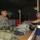 A U.S. Army soldier wearing ACU (Army Combat Uniform) with the 82nd Airborne Division patch and name tape 'LEABLE' is at a counter interacting with a civilian worker wearing a blue safety vest with yellow reflective striping. The setting appears to be a military equipment turn-in or supply facility, with piles of military gear, clothing, and bags on the counter between them. The background shows additional personnel and storage containers in a warehouse-like structure.