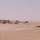 A desert landscape showing what appears to be an early-stage construction site with skeletal wooden or metal frame structures partially erected in sandy terrain. A light-colored vehicle is parked near the structures, and scattered debris and mounds of earth are visible across the flat, arid ground. The scene suggests preliminary building activity in a remote desert location.