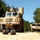 A military convoy on a gravel road consisting of a large MRAP (Mine-Resistant Ambush Protected) vehicle in the foreground equipped with a mounted weapon system and large sensor/antenna array, accompanied by a Humvee in the background. A small unmanned ground vehicle (UGV/robot) is positioned in front of the MRAP, likely being used for route clearance or reconnaissance. The scene appears to be a training exercise in a wooded area.