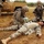 A military field medical training exercise showing U.S. Army soldiers performing casualty care on a simulated wounded soldier lying on the ground. Two soldiers are actively treating the downed soldier, one wearing blue medical gloves and applying treatment, while another holds what appears to be an IV bag. A military vehicle (HMMWV/MRAP) is visible in the background with a caution warning about speed.