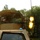 A soldier in camouflage gear is operating a mounted machine gun from the turret of a military vehicle, with a visible muzzle flash indicating the weapon is being fired. The vehicle appears to be a tan/desert-colored military truck with camouflage netting draped over the turret area. The setting is a wooded area, and text on the vehicle windshield reads 'WOLVERINE 51P'.