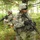 Two U.S. Army soldiers in full combat gear are moving through a wooded area with dense undergrowth. Both are wearing Army Combat Uniform (ACU) digital camouflage pattern and carrying M4 carbine rifles. The foreground soldier wears a PASGT/ACH helmet with an attached device and displays a U.S. flag patch on the left shoulder.