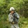 A soldier in US Army ACU (Army Combat Uniform) digital camouflage pattern is moving at a jog or run through a wooded training area. The soldier is fully equipped with combat gear including a helmet, body armor, and a rifle with what appears to be a MILES (Multiple Integrated Laser Engagement System) training device attached, indicated by the yellow component visible on the weapon. The setting and training equipment suggest this is a field training exercise rather than a combat deployment.