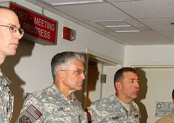 Four individuals stand in a corridor outside a meeting room, three of whom are wearing US Army ACU (Army Combat Uniform) camouflage uniforms. A illuminated red sign above the door reads 'QUIET - MEETING IN PROGRESS'. The fourth individual is a civilian wearing a plaid shirt and holding a notepad.