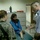 A medical or clinical consultation scene showing a woman in a blue garment and dark hijab seated on an examination table. Two individuals in U.S. Army camouflage uniforms are present alongside a civilian male who appears to be explaining or demonstrating something with an open-hand gesture. The setting appears to be a medical facility, possibly in a Middle Eastern location based on visible Arabic text on posted materials.
