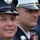 The image shows three uniformed firefighters, likely from New York, standing in formation. The foreground subject is in sharp focus wearing a dark navy dress uniform cap with a badge marked 'N.Y.', while two other firefighters are visible but out of focus in the background. The setting appears to be a formal or ceremonial occasion based on their dress uniforms.