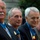 Four older men are seated together, each wearing the United States Medal of Honor on blue ribbons around their necks. They appear to be at a formal outdoor ceremony or commemorative event. A bronze statue or sculpture is partially visible in the background.