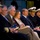 A group of approximately seven individuals are seated in a row at what appears to be a formal military or official ceremony. The group includes both uniformed naval officers wearing white peaked caps with gold insignia and civilians in business attire. Several individuals are holding printed programs or documents, suggesting they are attendees at a formal event.