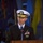 A senior U.S. Navy officer in dress uniform is speaking at a podium bearing the United States Navy Memorial seal. The officer is wearing a white-topped service dress blue uniform with multiple rows of ribbons, aviator wings, and flag officer insignia on his cap. The setting appears to be a formal military ceremony.