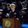 A senior U.S. Navy officer in dress blues is speaking at a podium bearing the United States Navy Memorial seal. The event appears to be a formal military ceremony, with numerous Navy personnel in dress uniforms visible in the background. The setting suggests an official commemorative or ceremonial occasion at the U.S. Navy Memorial.