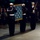 A formal military ceremony featuring U.S. Navy personnel in dress uniforms presenting or displaying a flag with white stars on a blue field with gold fringe, consistent with the Medal of Honor flag. Seated observers in the background include at least one individual wearing what appears to be a medal around their neck. The ceremony takes place on a stone or marble floor in a dimly lit venue.