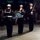 A formal military ceremony is taking place at night, featuring U.S. Navy personnel in dress uniforms. Two sailors are carrying a folded American flag with gold fringe, while a third stands at attention with a rifle. Seated observers in the background include individuals wearing military medals, suggesting a high-honor commemorative event.