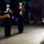 A formal U.S. Navy military ceremony is taking place at night in what appears to be an outdoor or semi-outdoor venue. Enlisted sailors in dress blues with white covers are performing a flag folding ceremony, while a senior officer in dress uniform stands at attention to the right. Seated observers in formal attire are visible in the background.