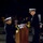 A formal U.S. Navy military ceremony taking place at night, likely at the Navy Memorial in Washington D.C. Two naval officers in dress uniforms are exchanging or presenting a folded flag or ceremonial item near a podium. An audience is visible in the background observing the proceedings.