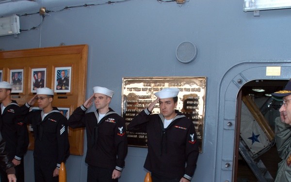 A formal military ceremony is taking place aboard what appears to be a U.S. Navy vessel, likely a museum ship or active warship. Several Navy sailors in dress uniforms are rendering salutes to an elderly veteran wearing a garrison cap and a flight jacket, while an officer in a green flight jacket observes from a hatchway doorway. A brass commemorative plaque is mounted on the gray bulkhead wall behind the sailors.