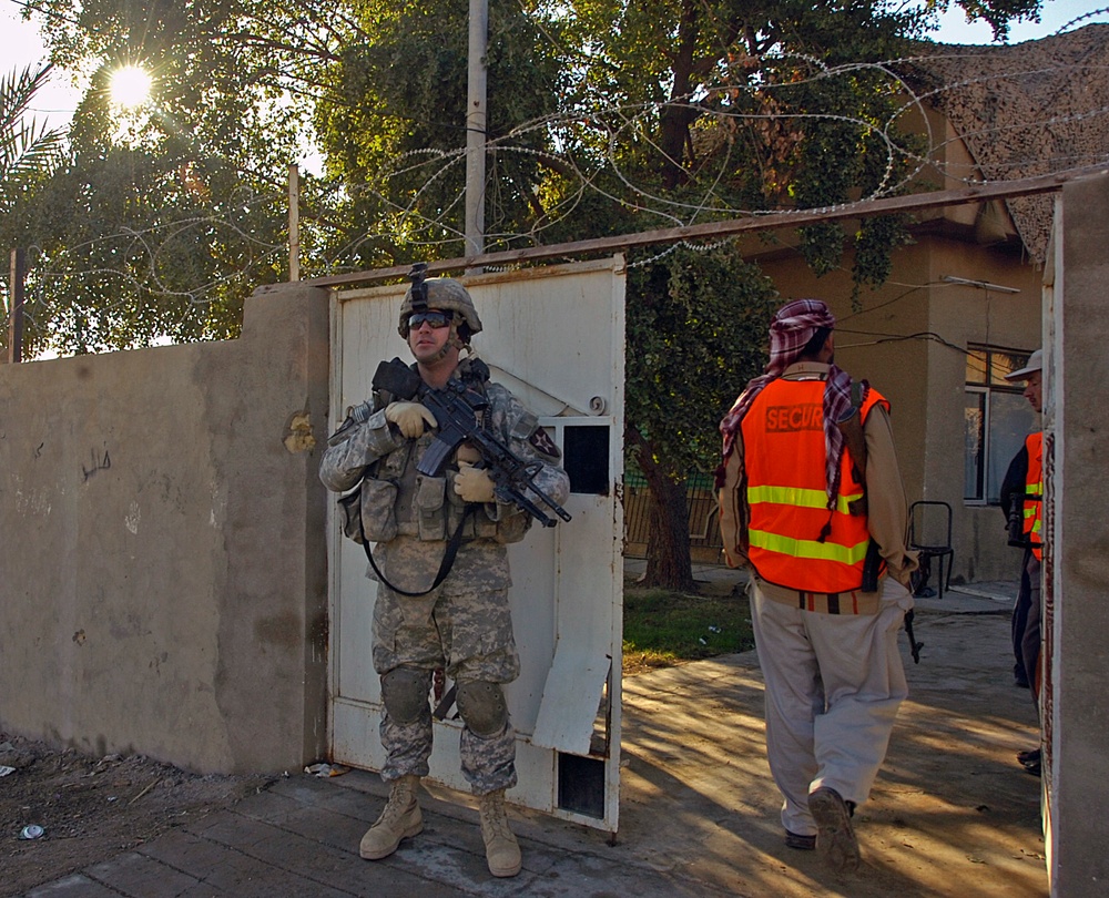 A U.S. Army soldier in full combat gear stands guard at a gated compound entrance secured with razor wire. A local security personnel wearing an orange high-visibility vest marked 'SECUR[ITY]' and a traditional red-and-white keffiyeh is walking through the gate. The setting appears to be a secured facility in the Middle East, likely Iraq, based on the architecture, vegetation, and Arabic writing visible on the wall.