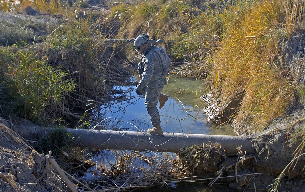 A soldier in US Army digital camouflage (ACU pattern) uniform and helmet is carefully crossing a water-filled irrigation canal or ditch by balancing on a narrow pipe or log. The surrounding terrain features dry scrub vegetation, reeds, and muddy banks consistent with an arid or semi-arid environment. The image appears to document a military patrol or reconnaissance operation in a rural area.