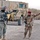 Sappers clear dangerous route essential to Operation Raider Harvest Two armed soldiers are coordinating at what appears to be a military checkpoint or base entrance. One soldier wears US Army ACU (Army Combat Uniform) with a 2nd Infantry Division patch, while the other wears desert camouflage with a red beret consistent with Iraqi Army military police. A large armored military vehicle (MRAP-type) is visible in the background near a concrete blast wall with Arabic graffiti.