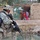 Sappers clear dangerous route essential to Operation Raider Harvest A U.S. Army soldier in full combat gear patrols near a small waterway in what appears to be a rural Middle Eastern village. In the background, a woman and several children stand near the entrance of a mud-brick compound, observing the soldier. The scene depicts a civil-military interaction during what appears to be a patrol or area security operation.
