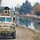 Sappers Clear Dangerous Route Essential to Operation Raider Harvest A tan/desert-colored MRAP (Mine-Resistant Ambush Protected) military vehicle is traveling along a narrow dirt road beside an irrigation canal in what appears to be a rural Middle Eastern or Central Asian setting. The vehicle is equipped with a gun turret covered by camouflage netting and multiple antennas. Civilians, including adults and children, are visible in the background walking along the same road.