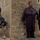Sappers clear dangerous route essential to Operation Raider Harvest The image shows a U.S. soldier in full combat gear taking a kneeling position at the corner of a brick building, while two civilian men in traditional robes stand nearby against the wall. Arabic graffiti is visible on the building to the left. The setting appears to be an urban or semi-rural area consistent with Middle Eastern architecture.