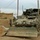 Sappers clear dangerous route essential to Operation Raider Harvest The image shows a heavily equipped military armored fighting vehicle, likely a Bradley IFV or similar tracked APC, fitted with a front-mounted bulldozer blade and slat/cage armor on its sides. The vehicle is positioned in what appears to be a Middle Eastern urban or semi-rural environment with mud-brick structures visible. The vehicle shows signs of heavy field use and modification, including additional equipment stowed on top.