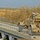 Sappers clear dangerous route essential to Operation Raider Harvest A sand/tan-colored military wheeled vehicle, likely an armored engineering or recovery vehicle, is positioned at the end of a concrete bridge with blue steel railings and support structure. The bridge appears to cross a waterway or low-lying area surrounded by tall dry reeds and scrubland. The setting appears to be a arid or semi-arid rural landscape, possibly in the Middle East.