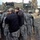 Sappers clear dangerous route essential to Operation Raider Harvest A group of U.S. Army soldiers in ACU (Army Combat Uniform) pattern camouflage are gathered in a huddle near a military armored vehicle covered with camouflage netting. One individual in the center is wearing civilian black clothing, possibly a contractor, journalist, or civilian official. The setting appears to be a forward operating base or staging area in a desert environment.