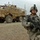 Sappers clear dangerous route essential to Operation Raider Harvest A U.S. Army soldier in full combat gear stands in the foreground holding an assault rifle, with a large Mine-Resistant Ambush Protected (MRAP) vehicle visible in the background. The soldier is wearing Army Combat Uniform (ACU) digital camouflage pattern, ballistic eyewear, a helmet with a mounted device, and tactical gloves. The setting appears to be a arid, rocky terrain consistent with Middle Eastern operational environments.