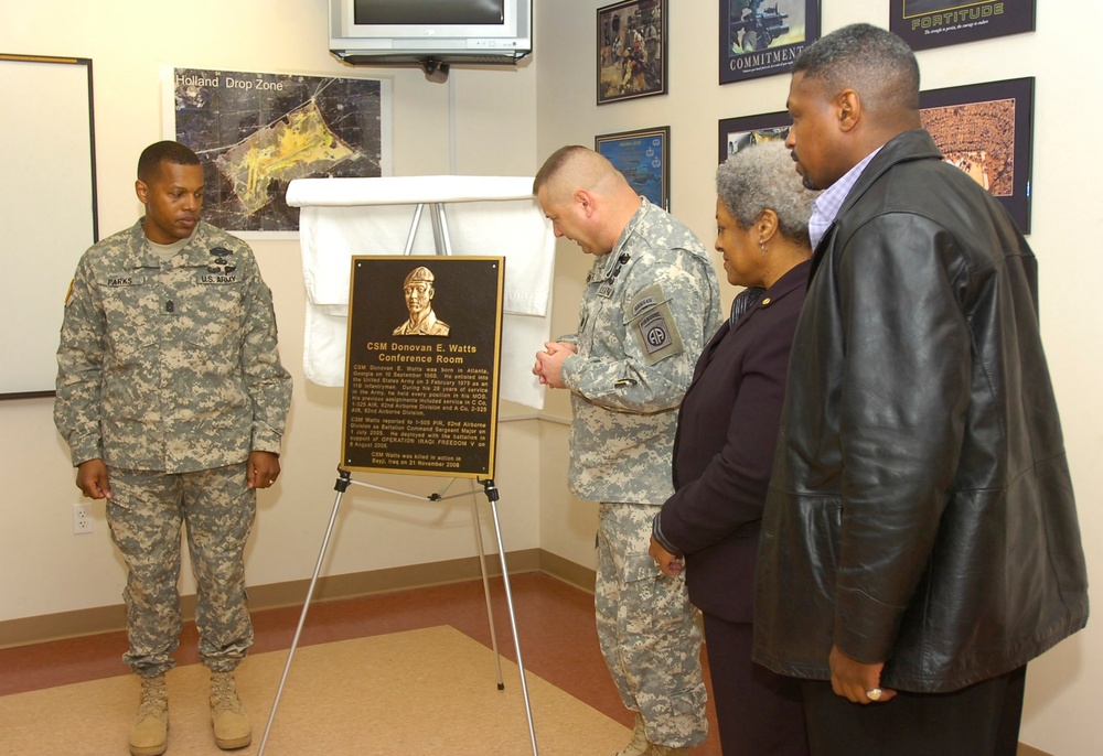 A military dedication ceremony is taking place in what appears to be a U.S. Army facility, where a bronze memorial plaque for 'CSM Donovan E. Watts Conference Room' is being unveiled on an easel. Two soldiers in Army Combat Uniforms and two civilians are gathered around the plaque, with one soldier bowing his head in a moment of reflection or prayer. The setting includes military-themed wall decorations, a map labeled 'Holland Drop Zone,' and motivational posters.