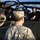 The image shows U.S. Army soldiers in ACU (Army Combat Uniform) interacting with a civilian near what appears to be an AH-64 Apache attack helicopter. A soldier with the name tape 'PENNINGTON' is visible from behind, facing a civilian in a black jacket who appears to be explaining or demonstrating something inside the cockpit. The helicopter bears a red number '1' insignia on its fuselage.