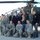 A group of approximately 13 individuals pose together in front of a military helicopter, likely an AH-64 Apache, on a tarmac or airfield. The group includes both uniformed U.S. Army soldiers in ACU (Army Combat Uniform) camouflage and civilians wearing casual clothing, several of whom appear to have USO badges or lanyards. The setting suggests a USO tour visit to a military installation, likely overseas.
