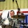 A U.S. Army soldier in ACU (Army Combat Uniform) is playing drums on a stage, smiling broadly while holding drumsticks mid-strike. The performance appears to be taking place at a military venue, likely a morale or entertainment event, evidenced by patriotic red, white, and blue decorations in the background. The drum kit includes visible cymbals branded 'CBI' and multiple microphones positioned around the kit.