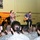 A USO concert performance taking place at what appears to be a military base, with a band performing on a small stage for an audience of uniformed military personnel. The band includes a vocalist, guitarist, drummer, and at least one other musician. A colorful painted backdrop with orange and red tones decorates the stage area.