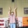 A male performer stands at a microphone stand on a stage with both arms raised above his head, appearing to be singing or performing. The venue is decorated with American patriotic imagery, military unit posters, and camouflage netting, suggesting a USO-style military entertainment event. A drum kit and other band equipment are visible in the background.