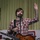A man with brown hair and a beard is performing on stage with an acoustic-electric guitar, wearing a red and white plaid shirt. He is standing at a microphone with one hand raised, appearing to address or wave to an audience. The setting appears to be an informal indoor venue with a green corrugated metal wall backdrop.